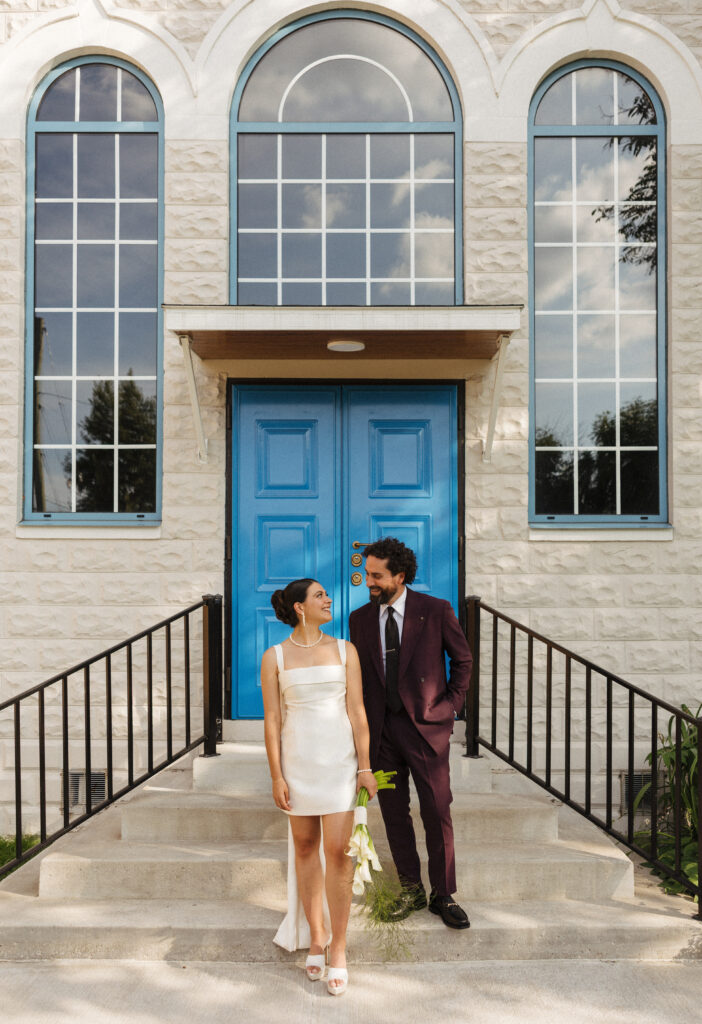 bride and groom portraits in front of white church with blue doors, groom in purple suit, bride in white mini dress